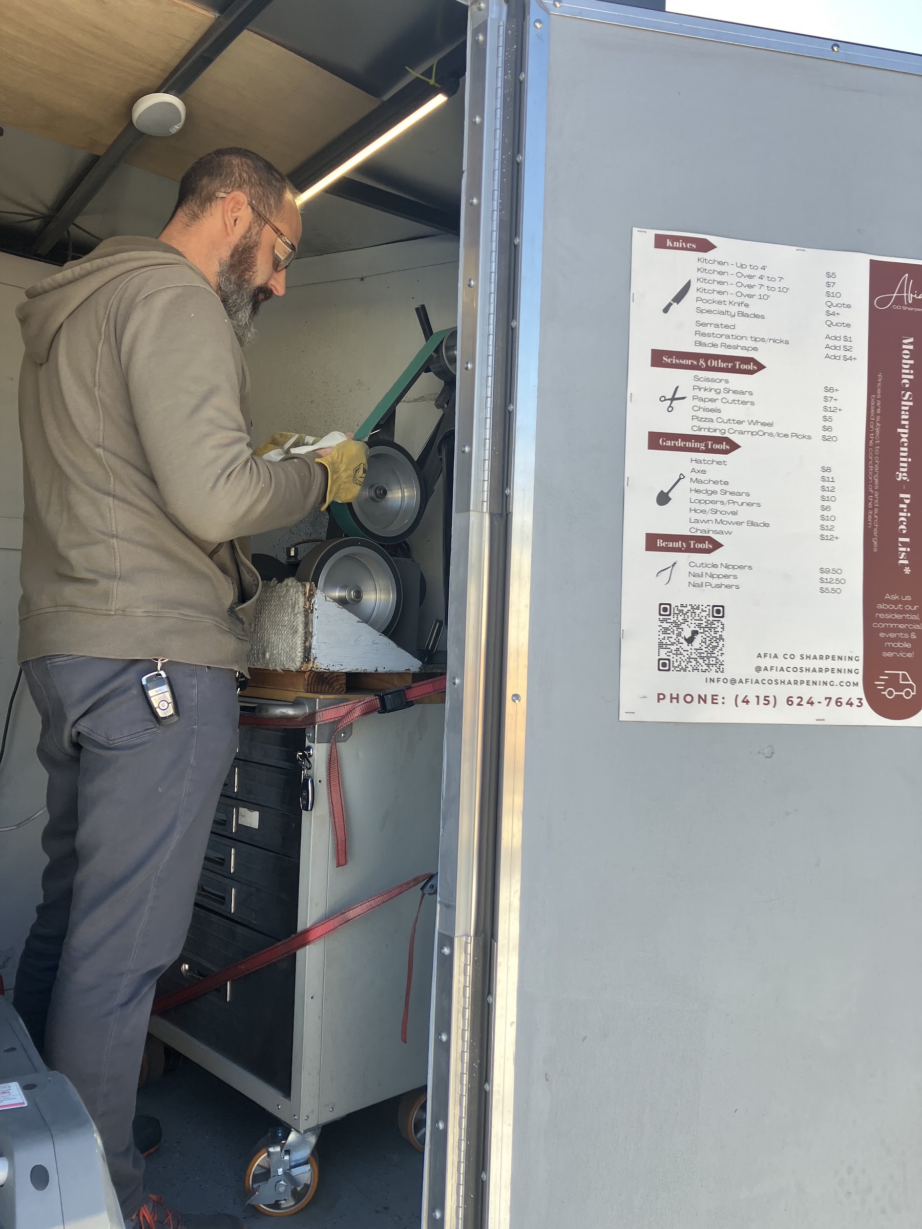 Erik sharpening inside the mobile trailer beside Afia's printed price list
